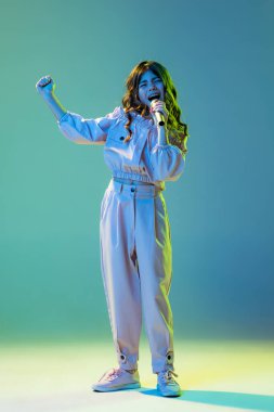 Musician, singer. Beautiful fashionable little girl, kid posing isolated over cyan color background in white neon light. Concept of children emotions, fashion, beauty, back to school and ad