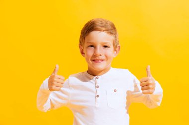 Baş parmaklar yukarı, güzel, güzel işaret. Sevimli küçük bir çocuğun portresi, parlak sarı arka planda izole edilmiş mutlu bir çocuk. Çocuk duyguları, yüz ifadesi, moda ve reklam kavramı. Mutlu görünüyor, mutlu.