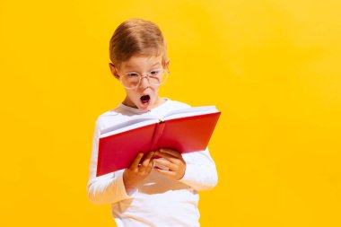 New information. Portrait of cute little boy, emotional kid looking at book with surprise isolated on bright yellow background with. Concept of child emotions, facial expression, fashion and ad.