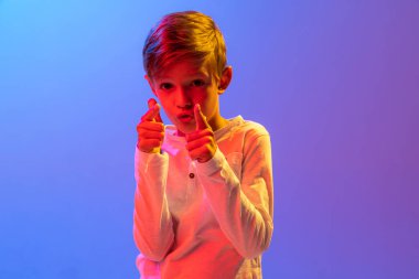 Stüdyo çekiminde şık küçük bir çocuk, neon ışıklarıyla izole edilmiş mavi-mor arka planda poz veriyor. Duyguların kavramı, yüz ifadesi, moda, hobiler, aktif yaşam tarzı ve reklam.