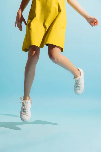 Lightness movements. Closeup slender female legs in summer dress and white sneakers hover above the floor isolated on blue background. Concept of beauty, art, fashion, youth, health