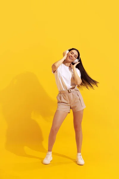 Soul music. Young charming girl with long dark hair in shorts and t-shirt isolated on bright yellow background. Concept of beauty, art, fashion, youth, health. Copyspace for ad