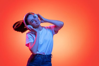 Müziği. Uzun siyah saçlı, kot pantolonlu ve tişörtlü, neonlu turuncu arka planda izole bir kızın stüdyo çekimi. Güzellik, sanat, moda, gençlik, sağlık kavramı. Reklam için kopyalama alanı