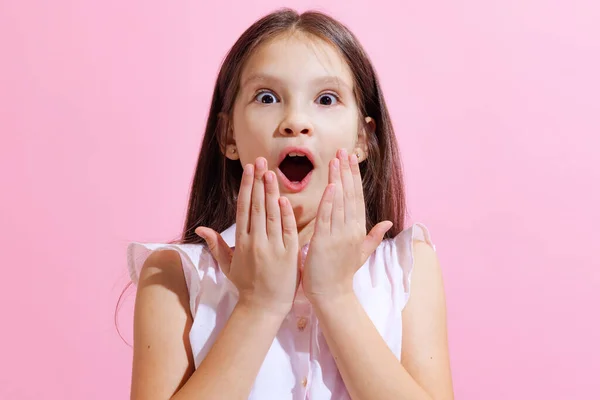 Wow, shock. Surprised little charming girl, kid looking at camera with open mouth isolated on pink background. Concept of children emotions, fashion, beauty, school and ad concept.