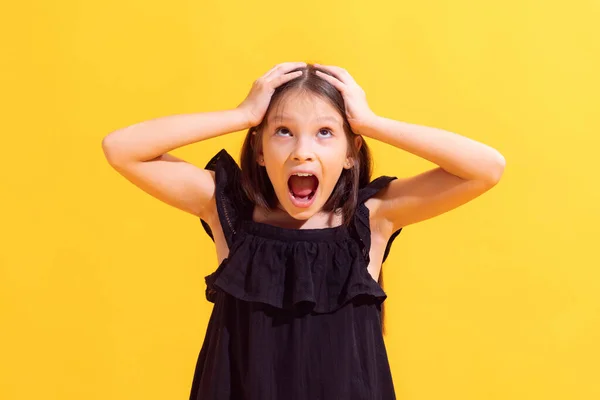 Irritation, anger and shock. Shouting little girl, kid wearing black dress isolated on yellow background. Concept of facial expressions, fashion, beauty, school and ad.