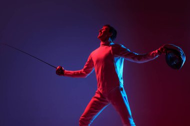Winner emotions. Portrait of young tall male fencer in fencing costume and mask holding smallsword isolated on purple background in neon. Copyspace for ad.