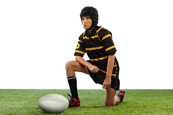 Junior athlete in sports uniform and helmet. One sportive boy, kid, male rugby player training with ball isolated on white background with grass floooring. Sport, team, studying, skills concept