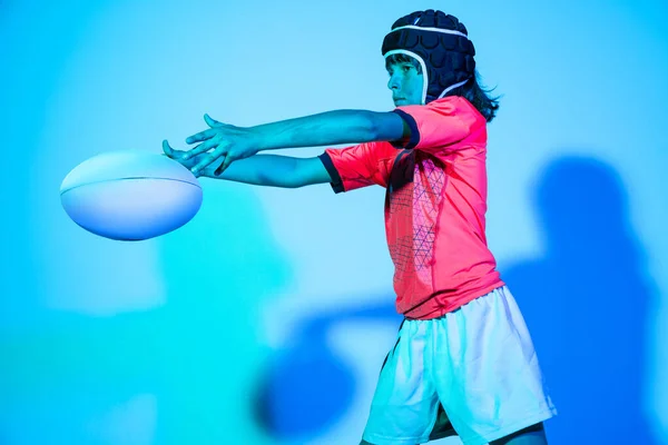 Portrait of junior male rugby player in sports uniform psoing with ball isolated on blue background in neon light. Sport, team, studying, skills concept