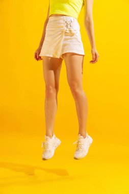 In jump. Slender female legs in white short skirt and sneakers hover above the floor isolated on yellow background. Concept of beauty, art, fashion, youth, healthy lifestyle.