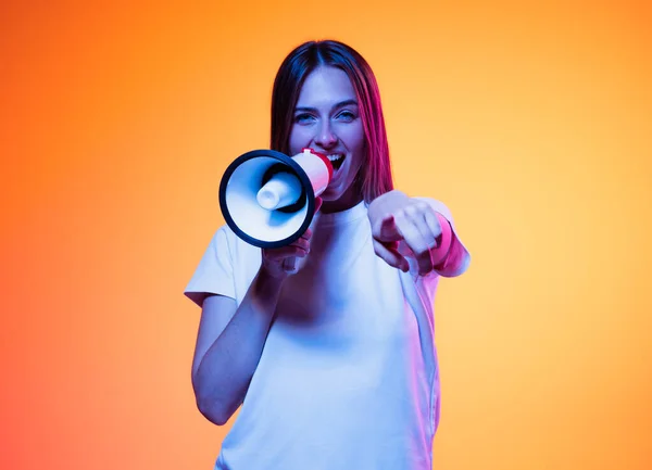 Shouting at megaphone. Young emotional girl, student in white t-shirt isolated on orange color background in neon light. Concept of info, propaganda, news