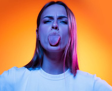 Showing a tongue. Closeup face of young attractive girl grimaces at camera isolated on orange color background in neon light. Concept of beauty, art, fashion, youth, sales and ad, education