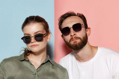 Serious people. Closeup young bearded man and beautiful girl in sunglasses looking at camera posing isolated over blue-pink background. Emotions, youth, facial expressions