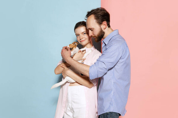 Hugs. Young couple in love with cute puppy posing isolated over blue-pink background. Emotions, love, care, support, relationship concept