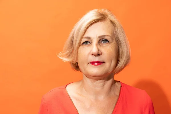 Calm. Half-length portrait of beautiful middle age woman with blond hair posing isolated on orange color background. Concept of natural beauty, ages, fashion, elder generation and ad
