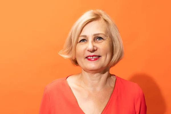 Smiling, happy. Half-length portrait of beautiful middle age woman with blond hair posing isolated on orange color background. Concept of natural beauty, ages, fashion, elder generation and ad