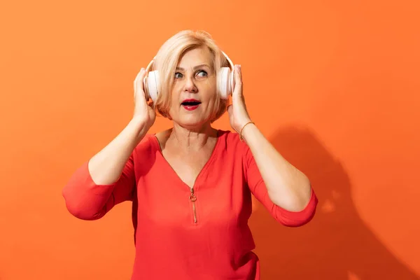 Terrible noise. Shocked middle age woman with blond hair in headphones isolated on orange color background. Concept of natural beauty, ages, fashion, elder generation and ad