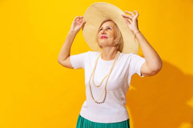 On vacation. Happy fashionable senior woman in summer style outfit and hat isolated on bright yellow background. Concept of beauty, emotions, vacation, travel, ad and sales