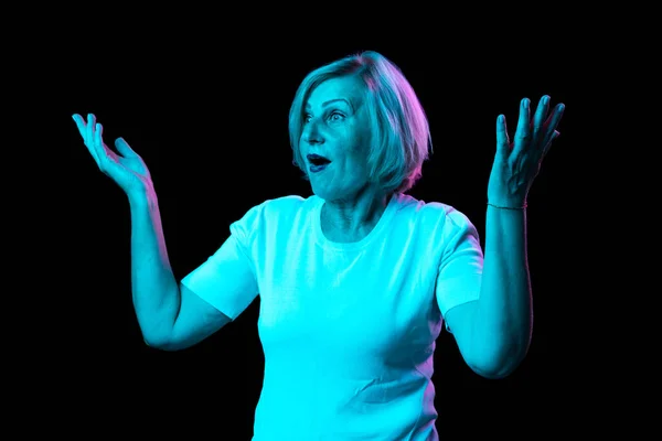 Wow, shock. Surprised senior woman with blond hair raising hands up in surprise isolated over dark background in neon light. Concept of natural beauty, ages, fashion, elder generation and ad