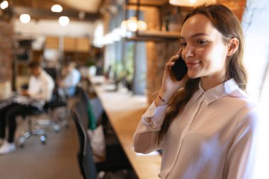 Phone conversation. Young charming smiling woman at loft style office with group of colleagues in background. Concept of success, business, career, achievements. looks happy, excited