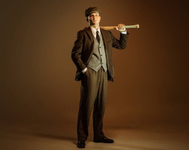 Retro style portrait of young man in image of english gangster, businessman wearing suit and cap standing isolated over dark vintage background. Concept of business, personality, emotions, fashion