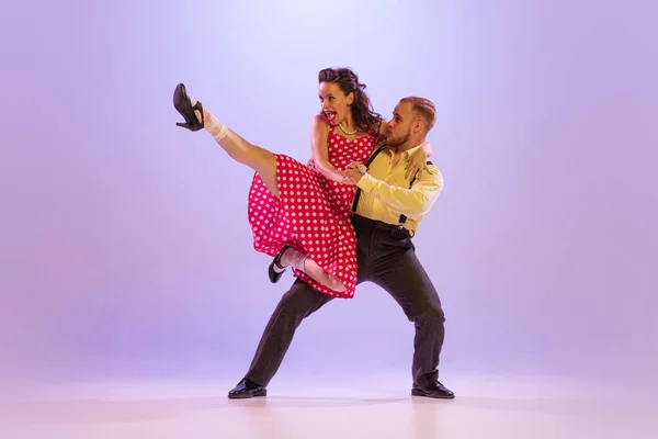 Lindy Hop. Active and emotional couple in colorful retro style costumes dancing incendiary dances isolated on purple background in neon. Actors in motion and action. Concept of art, 60s, 70s culture