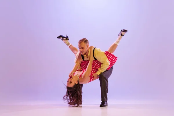 Love. Beautiful girl and man in colorful retro style costumes dancing incendiary dances isolated on lilac color background in neon light. Actors in motion and action. Concept of art, 60s, 70s culture