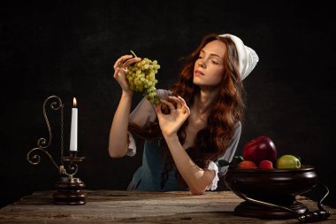 Tasting grapes. Young beautiful redhair girl with long curly hair like girl of renaissance eras isolated on dark background. Comparison of eras, beauty, history, art, creativity.