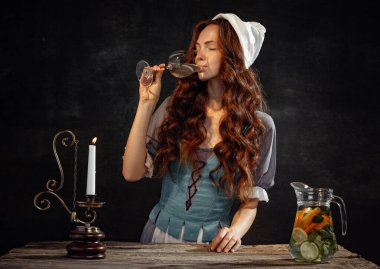 Tasting wine. Vintage portrait of young adorable redhead girl in image of medieval person, peasant woman in renaissance style dress isolated on dark background. Comparison of eras, beauty