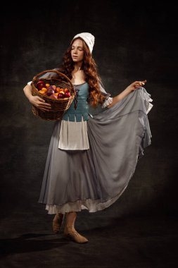Creative portrait of young beautiful redhead girl with long curly hair in image of medieval person in renaissance style dress isolated on dark background. Comparison of eras, beauty, history, art