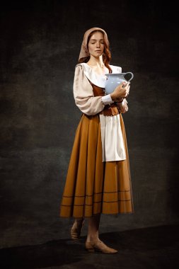 Vintage portrait of young adorable redhead girl in image of medieval person, peasant woman in renaissance style dress isolated on dark background. Comparison of eras, beauty, history, art, creativity.