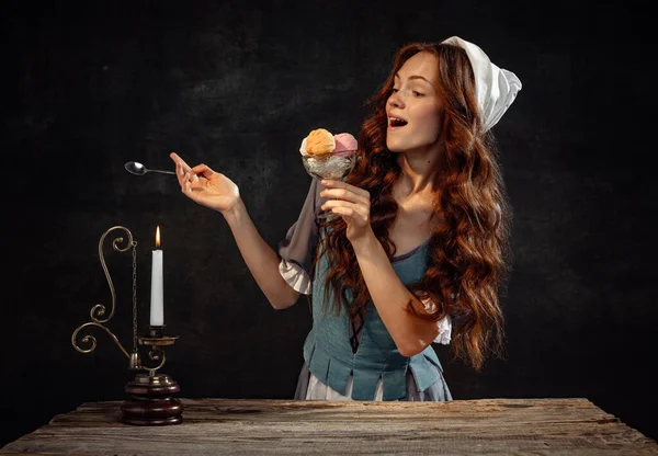 Ice-cream. Young beautiful redhair girl with long curly hair like girl of renaissance eras isolated on dark background. Comparison of eras, beauty, history, art, creativity.