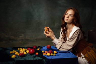 Fruits diet. Vintage style portrait of pretty redhead woman with long curly hair like girl of renaissance eras, lady-peasant isolated on dark background. Comparison of eras, beauty, history, art