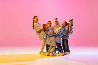 Modern choreography lesson. Dance group of happy, active little girls in t-shirts and jeans in action isolated on orange background in neon. Concept of music, fashion, art, childhood, hobby.