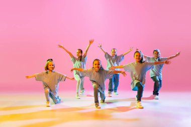 Joy, fun and happiness. Portrait of cheerful, active little girls, happy kids dancing isolated on orange background in neon light. Concept of music, fashion, art, childhood, hobby.