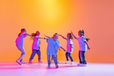Modern choreography lesson. Dance group of happy, active little girls in t-shirts and jeans in action isolated on orange background in neon. Concept of music, fashion, art, childhood, hobby.