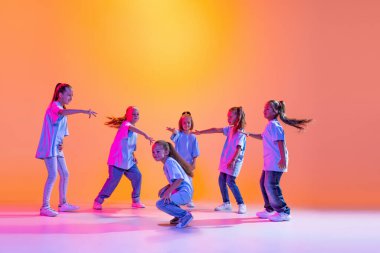 Joy, fun and happiness. Portrait of cheerful, active little girls, happy kids dancing isolated on orange background in neon light. Concept of music, fashion, art, childhood, hobby.