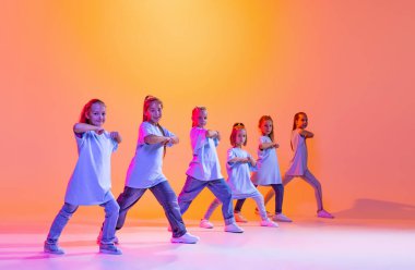 Joy, fun and happiness. Portrait of cheerful, active little girls, happy kids dancing isolated on orange background in neon light. Concept of music, fashion, art, childhood, hobby.