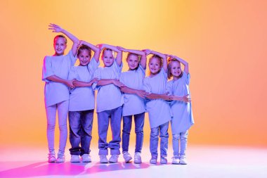 Joy, fun and happiness. Portrait of cheerful, active little girls, happy kids dancing isolated on orange background in neon light. Concept of music, fashion, art, childhood, hobby.