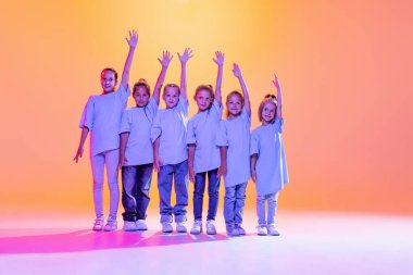 Modern choreography lesson. Dance group of happy, active little girls in t-shirts and jeans in action isolated on orange background in neon. Concept of music, fashion, art, childhood, hobby.