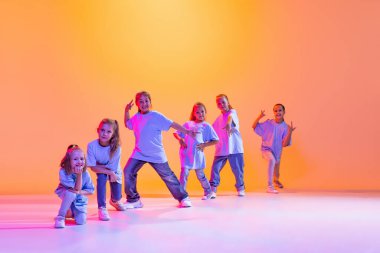Happy children dancing. Group of children, little girls in sportive casual style clothes dancing in choreography class isolated on orange background in purple neon light. Concept of music, fashion