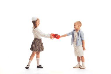 Portrait of beautiful little children, boy giving present box to girl isolated over white studio background. Concept of childhood, friendship, game, school, fun, education. Copy space for ad