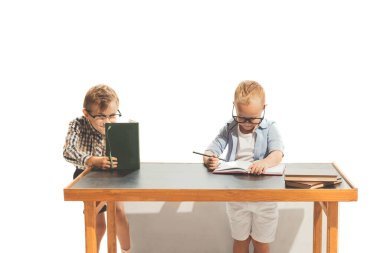 Portrait of two little boys, children, pupils sitting at desk, studying, doing homework isolated over white studio background. Concept of childhood, game, school, fun, education. Copy space for ad