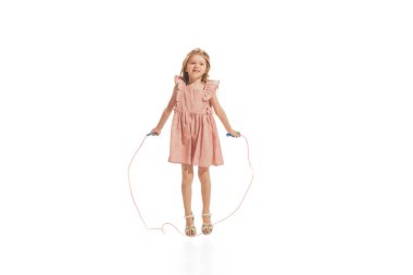 Portrait of beautiful little girl in stylish dress posing, jumping with rope, playing isolated over white studio background. Concept of childhood, game, fashion, fun, education. Copy space for ad