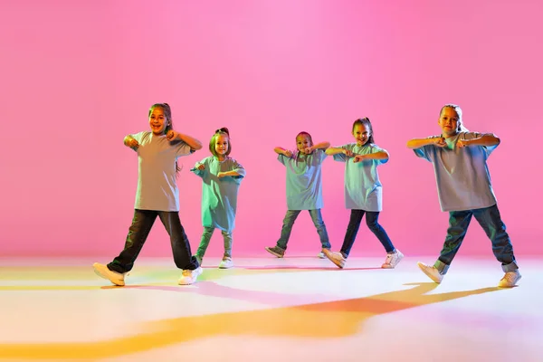 Happy children dancing. Group of children, little girls in sportive casual style clothes dancing in choreography class isolated on orange background in purple neon light. Concept of music, fashion