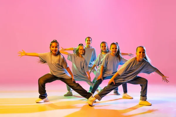 Hip-hop dance, street style. Happy children, little active girls in casual style clothes dancing isolated on orange background in purple neon light. Concept of music, fashion, art