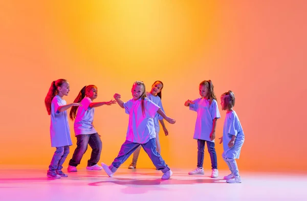 Happy children dancing. Group of children, little girls in sportive casual style clothes dancing in choreography class isolated on orange background in purple neon light. Concept of music, fashion