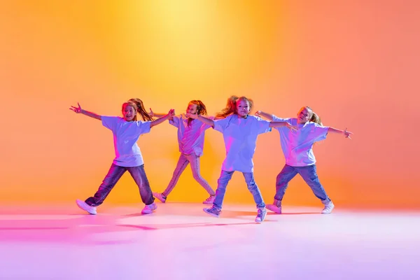 Happy children dancing. Group of children, little girls in sportive casual style clothes dancing in choreography class isolated on orange background in purple neon light. Concept of music, fashion