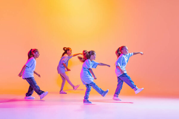 Hip-hop dance, street style. Happy children, little active girls in casual style clothes dancing isolated on orange background in purple neon light. Concept of music, fashion, art