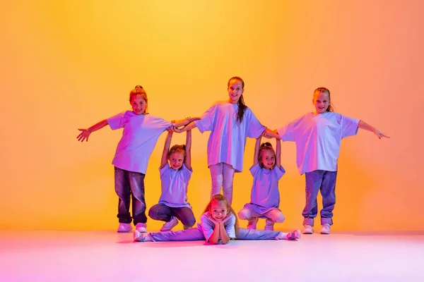 Modern choreography lesson. Dance group of happy, active little girls in t-shirts and jeans in action isolated on orange background in neon. Concept of music, fashion, art, childhood, hobby.