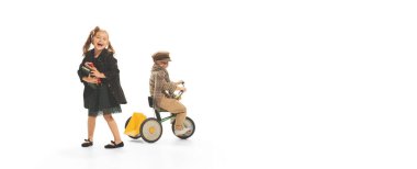 Portrait of cheerful girl with books playing with little boy on small bike isolated over white background. Retro fashion. Concept of childhood, game, school, fun, education. Copy space for ad